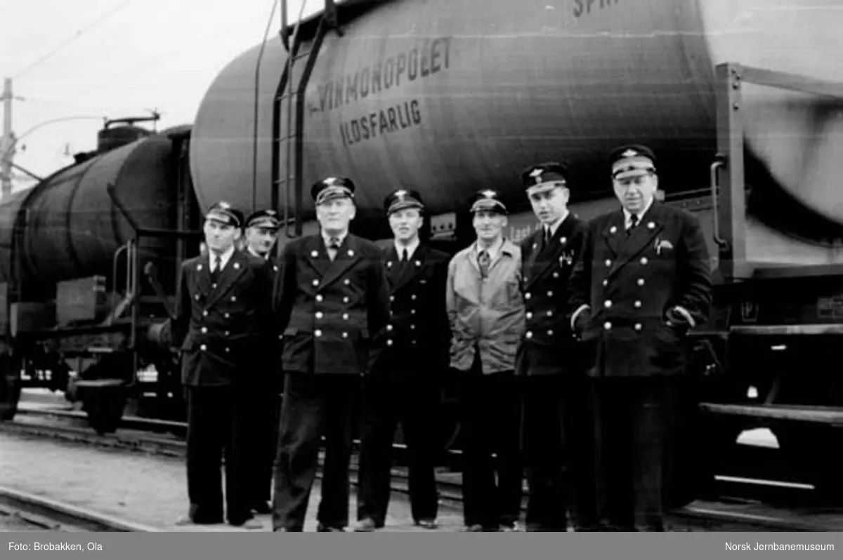 Gruppebilde av sju jernbanemenn på Hamar stasjon foran en sprit-tankvogn
