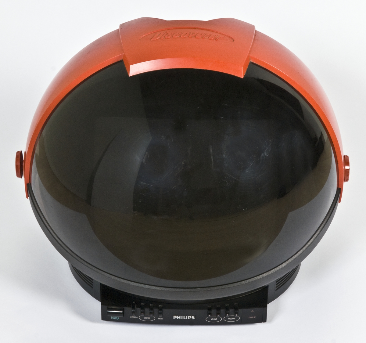 Tv-apparat utformet som en astronaut-hjelm med hjul bak og vippbart visir i gjennomsiktig mørk plast. Fjernkontroll med skyvedeksel følger med.