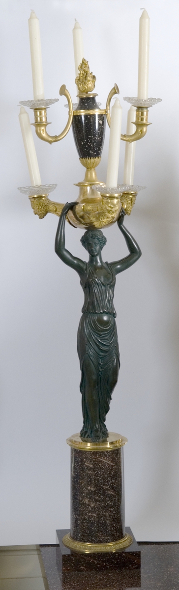 Kandelaber; porfyr, bronse forgylt og bronse mørk. Fotplate firkantet, sokkel rund, på den står kvinnefigur i klassisk gevant, hun bærer romersk lampe med tre lysholdere, over den urne med tre lysholdere.