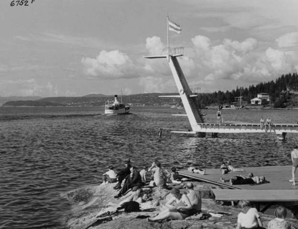 Strandliv med badegjester som soler seg på svaberget ved sjøen på Ingierstrand bad utenfor Oslo. Stupetårn og båt i bakgrunnen.