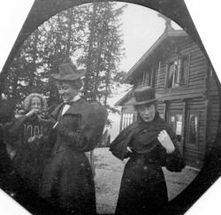 To kvinner og en ung pike foran tømmerhus i skogen, Golå, Sø