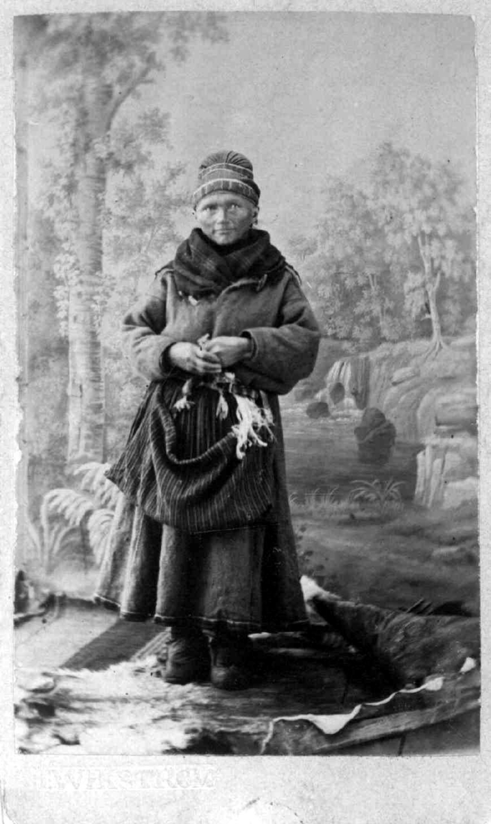 Kvinne i samedrakt fotografert i fotoatelier med malt skog i bakgrunnen. Antagelig Norrbotten.
