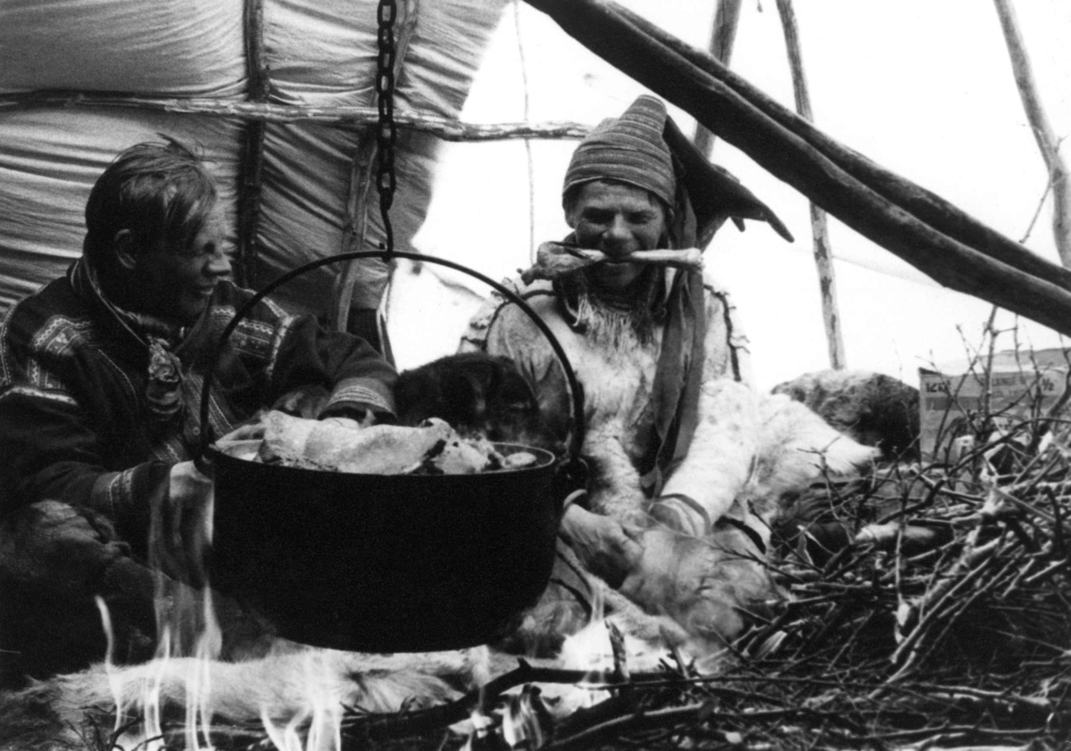 Kjøttkoking i telt. To menn sitter ved ildstedet med gryte over. Finnmark 1957.