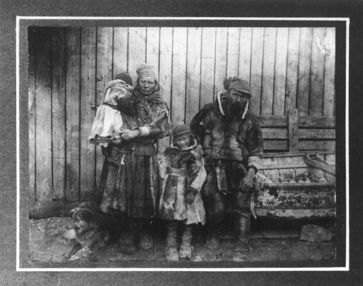 Samedrakter. To voksne og to barn foran en husvegg. Karesuando.