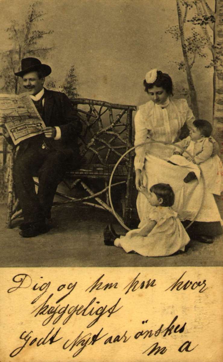 Postkort, Nyttårshilsen. Fotografi. Svart/hvitt. En mann sitter  på en benk og leser avisen. Til høyre på samme benk sitter en kvinne med et barn. et barn sitter på gulvet.