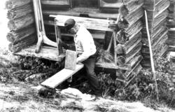 En mann demonstrerer rasping av frø utenfor et tømmerhus. Ha