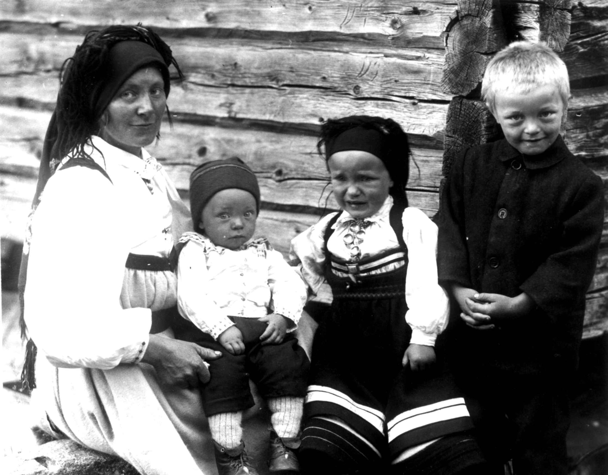 Kvinne- og barnedrakt, gruppeportrett, Valle, Setesdal, Aust-Agder, antatt 1924. Kvinne med tre barn sittende utendørs foran tømmervegg. Fra "De Schreinerske samlinger" (skal oppgis).