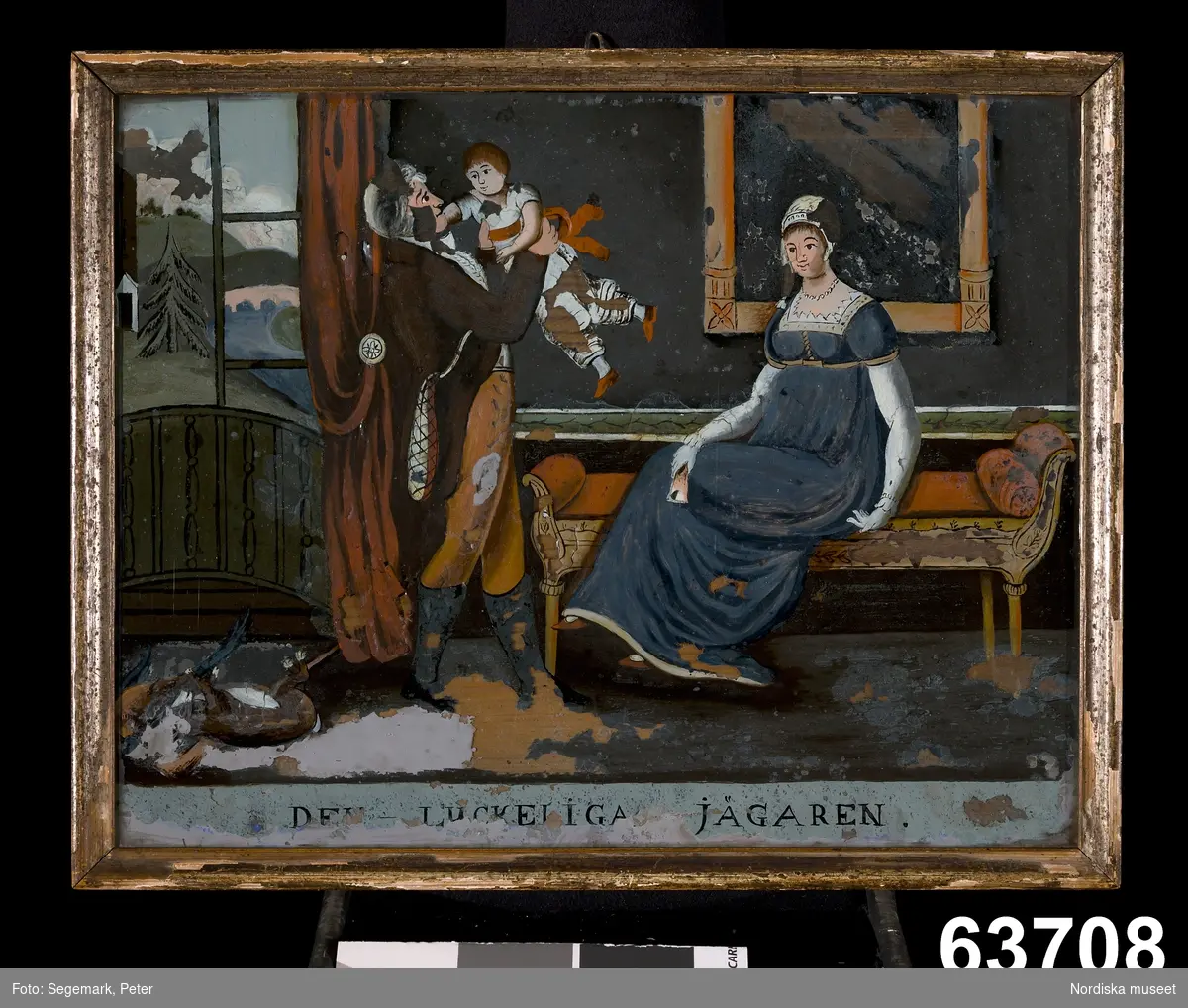 Interiör med man, kvinna och barn i Karl Johan-soffa, vid mannens fötter jaktbyte.
Djur