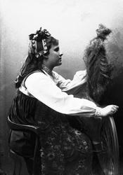 Studioportrett av kvinne i drakt og hodeplagg, som sitter ve