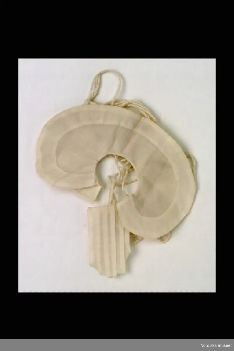 Inventering Sesam 1996-1999:
B 13 cm
Dockkrage av tunn vit bomull, rundad form, fem tvinnade bomullssnören att bla knyta under armarna för att fästa kragen.
Helena Carlsson 1997