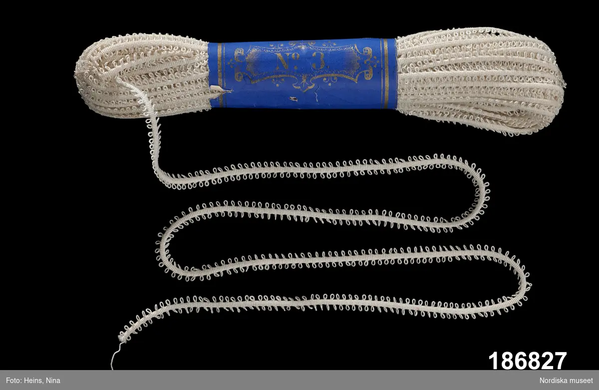 Spetsband.
Linne.
Maskingjort rundvävt smalt band med två jämna rader öglor. Metervara som ligger buntat med blå maggördel med "No. 3" i guldtryck.
/Berit Eldvik 2002