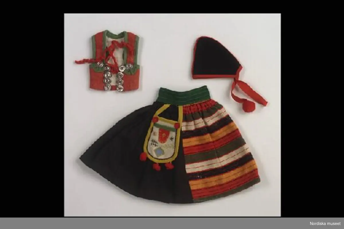 Inventering Sesam 1996-1999:
L 19, L 10, H 7 cm
Rättviksdräkt till docka.
a) kjol av svart ylle, linning av grönt ylle, i kjolen insytt ett randigt ylletyg i flera färger som skall föreställa ett förkläde. Väska i flera färger hängande i midjan.
b) livstycke av rött ylle, knappliknande snörmaljor av grå metall med hyskor under, rött snörband av yllegarn, kantad med grönt ylle.
c) hätta av svart kläde, i nacken ett par röda ylleband avslutade med röda bollar, en saknas.
Helena Carlsson 1997