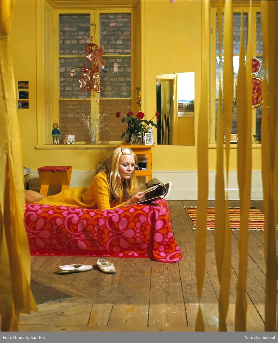 Kvinna ligger på sängen och läser tidning i rum med gula väggar.