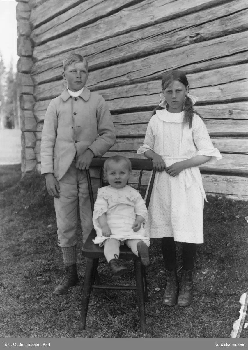 "Tre barn".  Pojke, flicka och ett litet barn i en stol utanför en timmerstuga. Transtrand socken, Dalarna.
