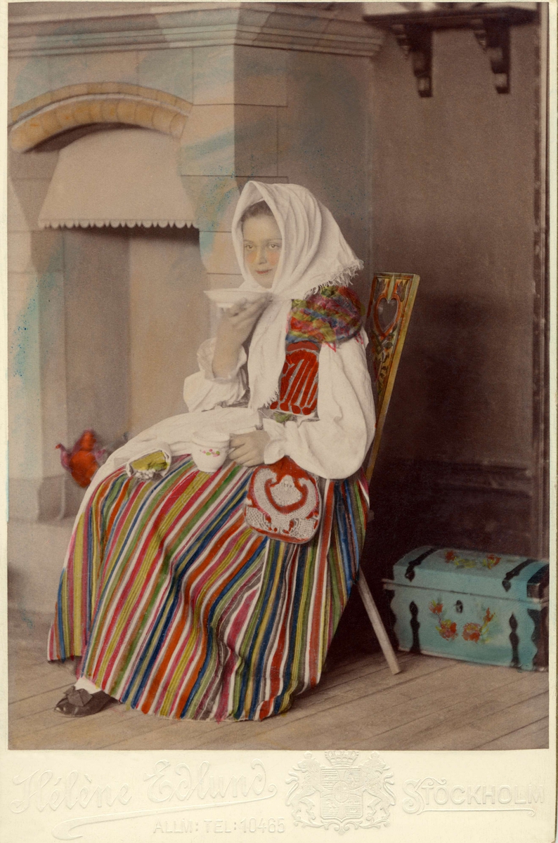 En kvinna klädd i dräkt från Östra härad i Småland sitter i ett kök och dricker kaffe.
