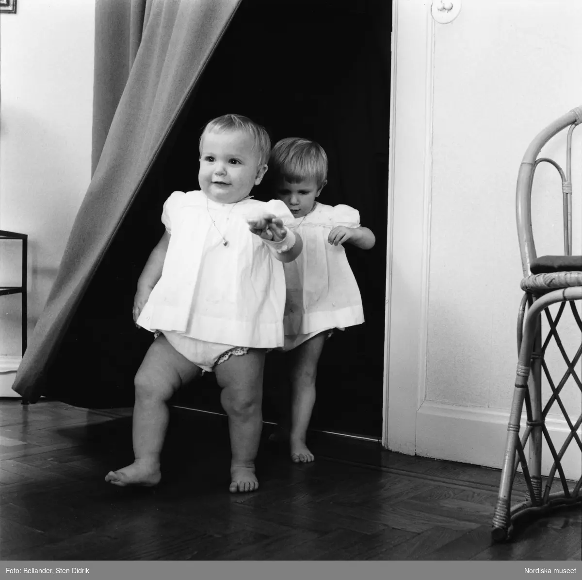 Barnporträtt. Två små flickor i ettårsåldern klädda i ljusa klänningar marscherar in i ett rum.