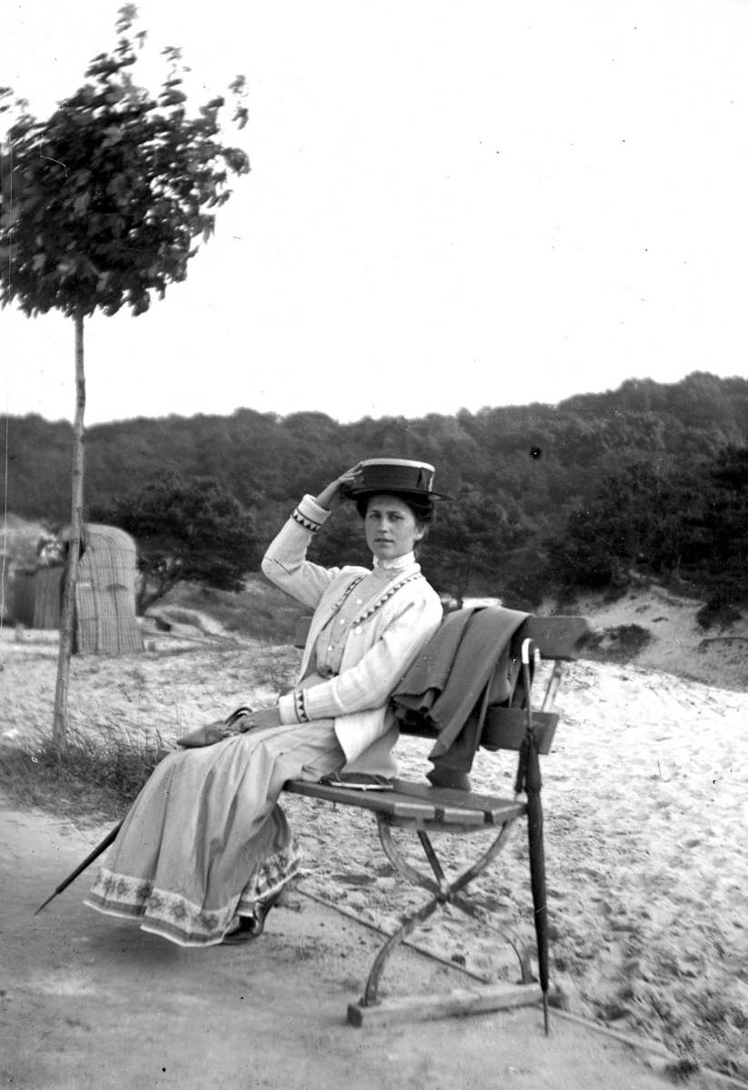 Porträtt av en kvinna sittande på en bänk i utkanten av ett dynlandskap. Till vänster skymtar en flätad badhytt.