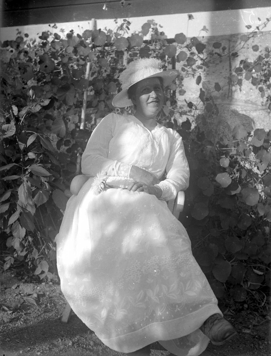 Porträtt av en kvinna i vit hatt och klänning. I bakgrunden en husgrund och slingerkrasse.