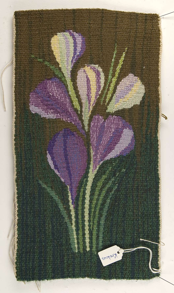 Bildväv föreställande lila krokus mot grön och brun bakgrund. Bilden är en flamskvävnad av ull och lin.
Fastknuten vid bildväven sitter en etikett med texten "Krokus Pris: 96:-"