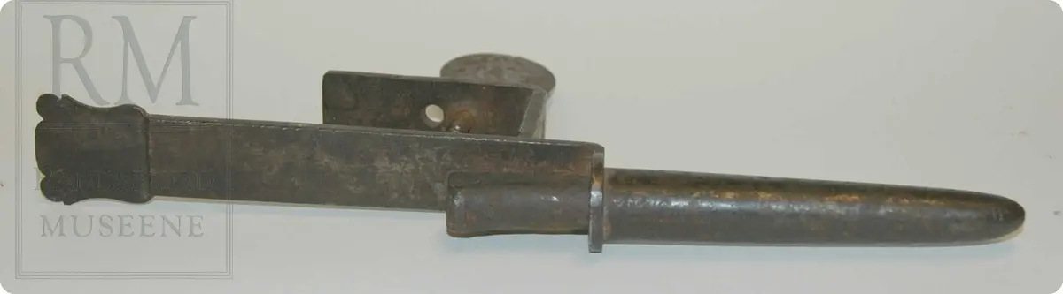 Avlangt jernstykke, en sylinderformet beholder i den ene enden, går opp til et fest på baksiden, framsiden er rektangelformet, med et lite "våpen" på toppen.
