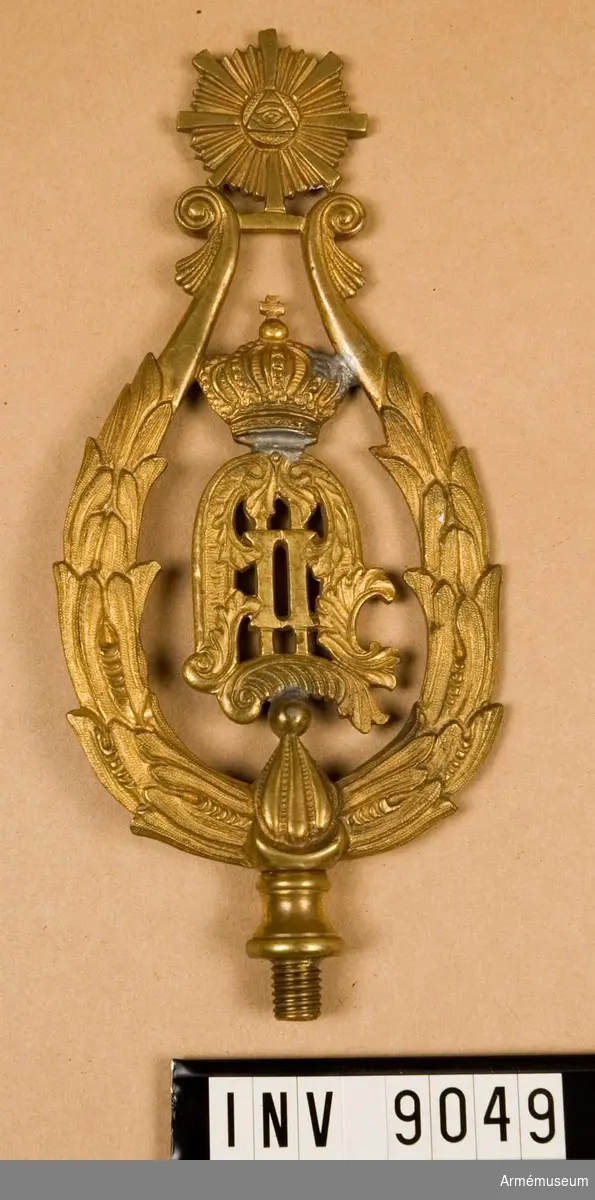 Av förgylld metall med konungens monogram O II omgivet av en krans, spetsformad. För fastsättning vid fanan en gängad skruv.