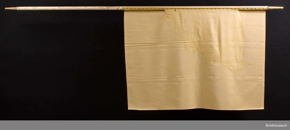 Duk av gult fyrskaftat fansiden. Fragment uppsydda på gult konstsiden. Fragmentet är från fanstången mot mitten 85 cm djupt. Stång 2.67 m lång av vitmålad furu. Doppsko saknas. Spets med Karl XV:s namnchiffer.