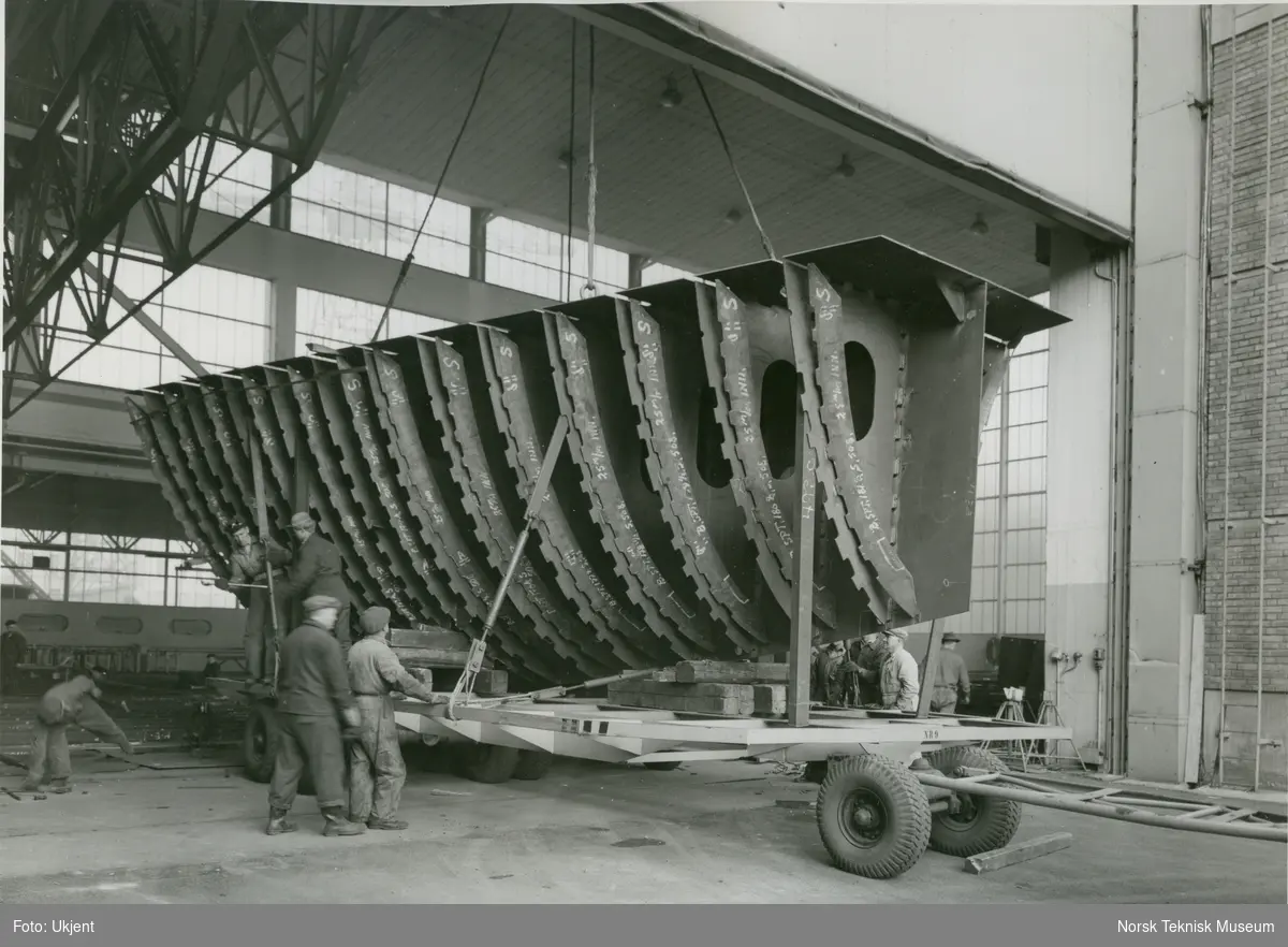 Arbeidere frakter en del fra båtbyggerhallen til lasteskipet M/S Baghdad, B/N 493c på Akers Mek. Verksted. Skipet ble levert i 1956 til Fred. Olsen & Co.