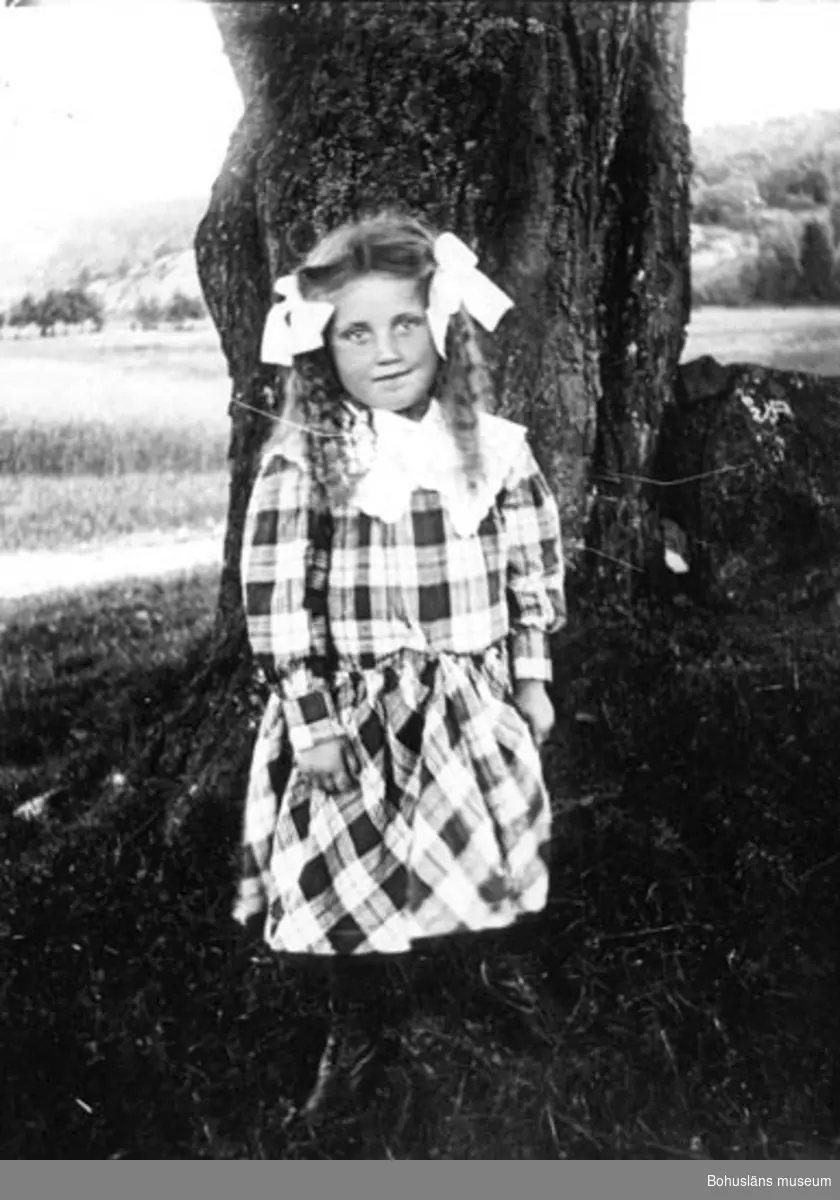 Flicka i rutig klänning, fotograferad utomhus framför en trästam.