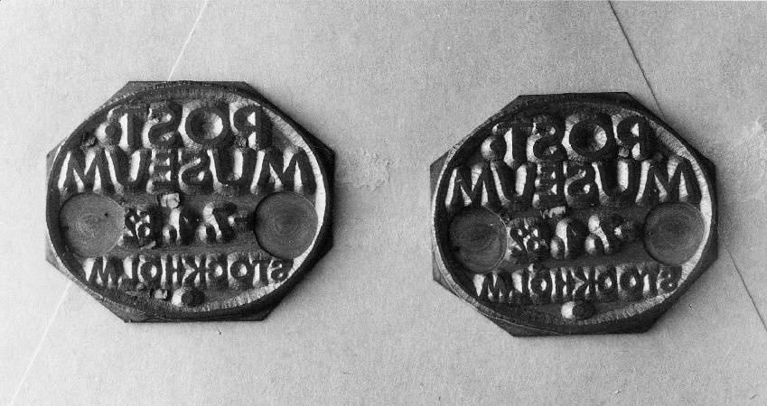 Kliché till metall- och gummistämpel använd i Postmuseum.
Åttkantig med ovalt stämpelavtryck (diam 30 mm) och spegelvänd text.