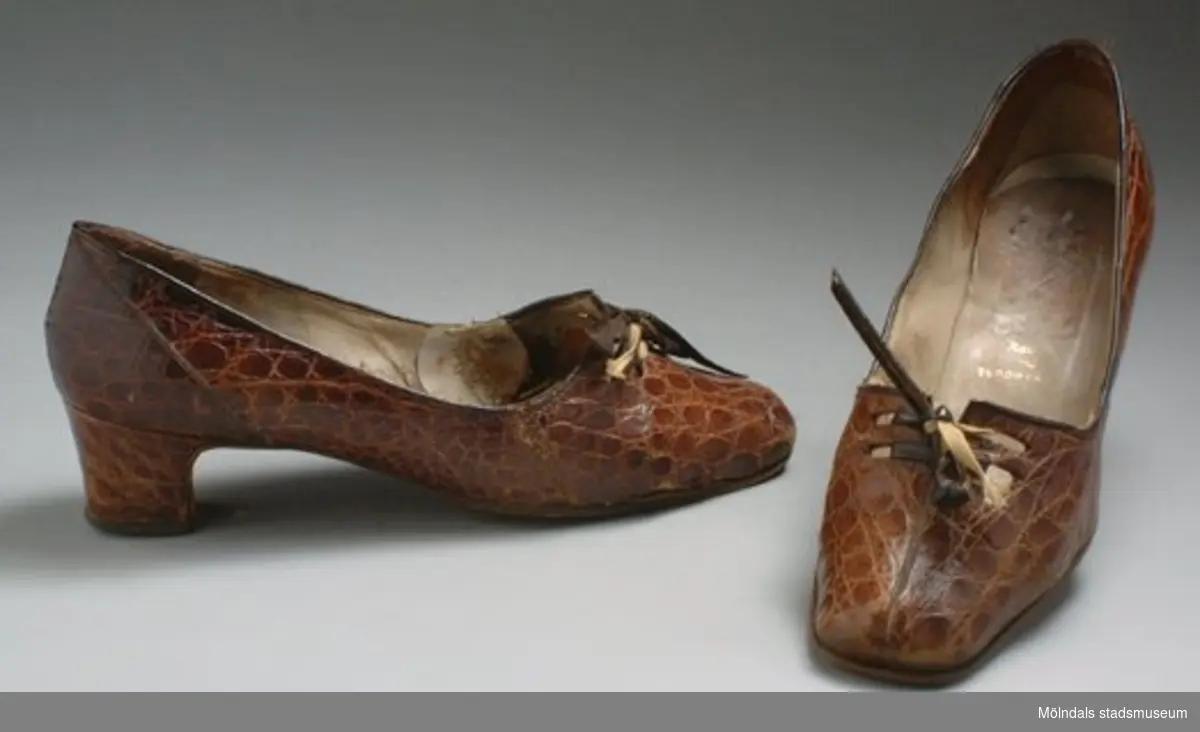 Brun sko i krokodilskinn med hög klack. Troligen inköpt i Innsbruck, Österrike.Slitna och lagade. Det finns tillhörande handväska: inv. MM03328.Brukade användas till klänning: inv. MM03323:1-4.
