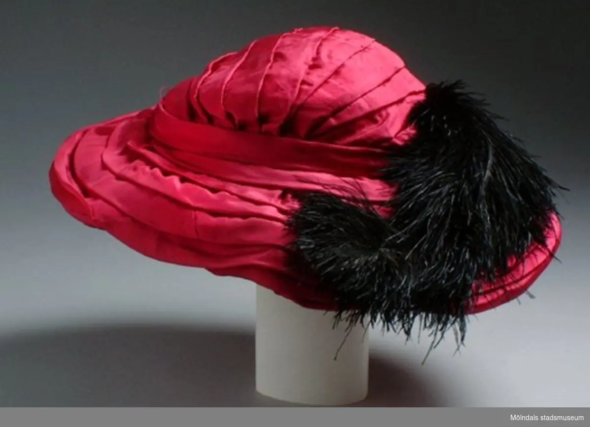 Cerise damhatt, något ovalformad med svart plym och tillhörande hårnålar.Handsydd av ägarens gudmor. Denna hatt låg i samma hattask inv.nr: 03404, som hatt med inv.nr: 03339.