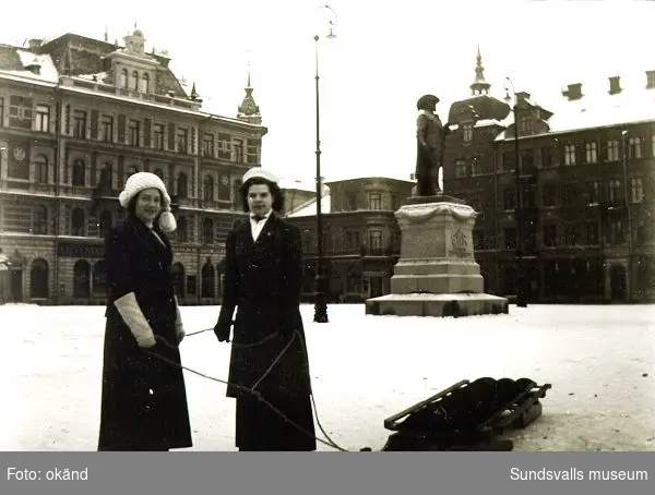 Två kvinnor med kälke på Stora torget i Sundsvall. I bakgrunden syns Harald Sörensen-Ringis staty av Gustav II Adolf, rest 1911.