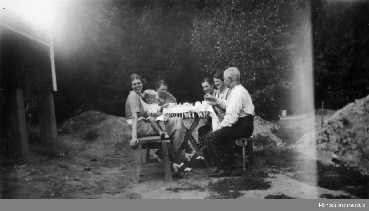 Kvinnor, män och ett barn vid ett dukat kaffebord utomhus. Bilden märkt: "Från Kållered". 1930-tal.