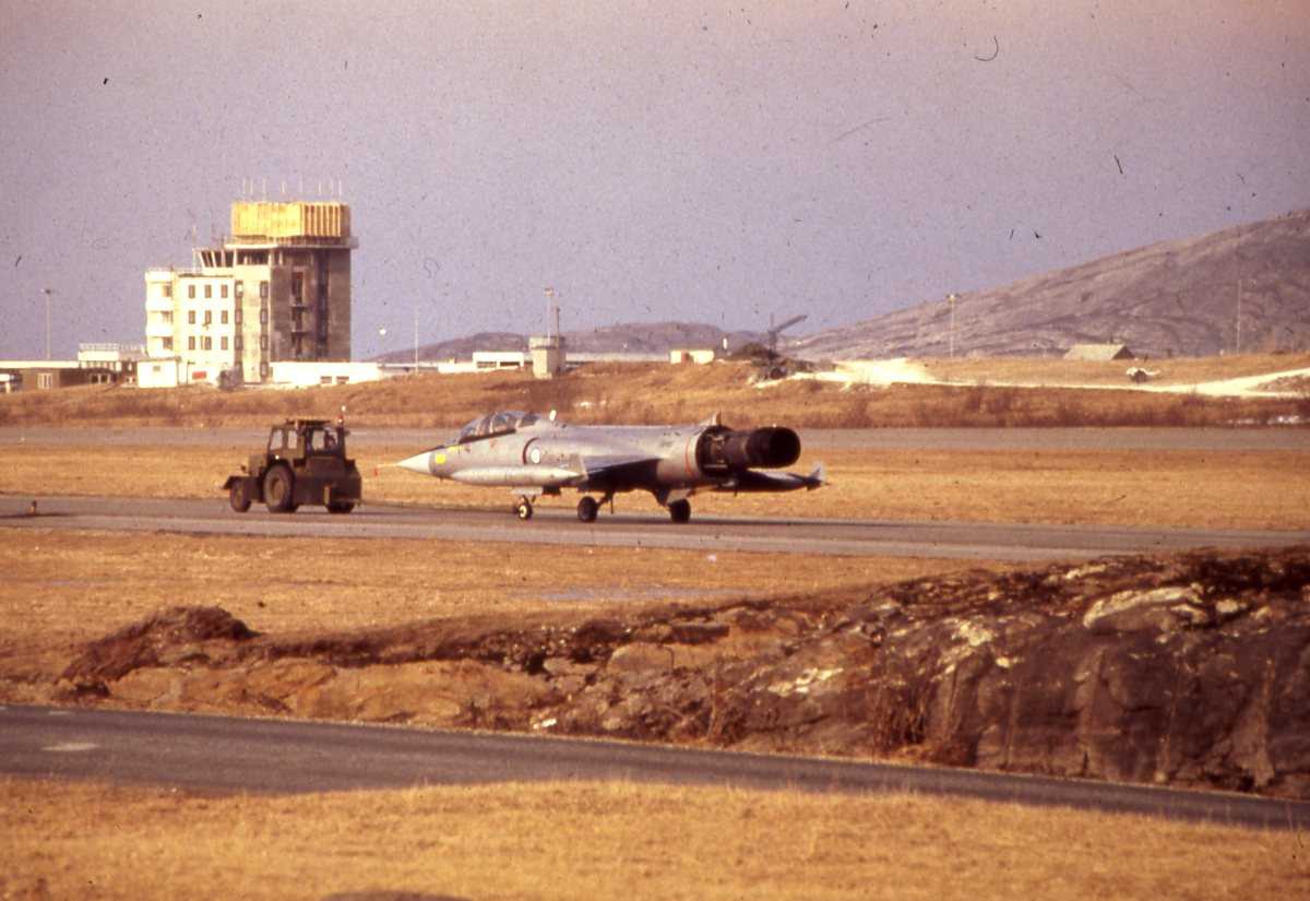 Norsk TF-104G Starfighter som slepes av et militært kjøretøy på Bodø hovedflystasjon. I bakgrunnen sees det nye kontrolltårnet som er i ferd med å bli bygget.