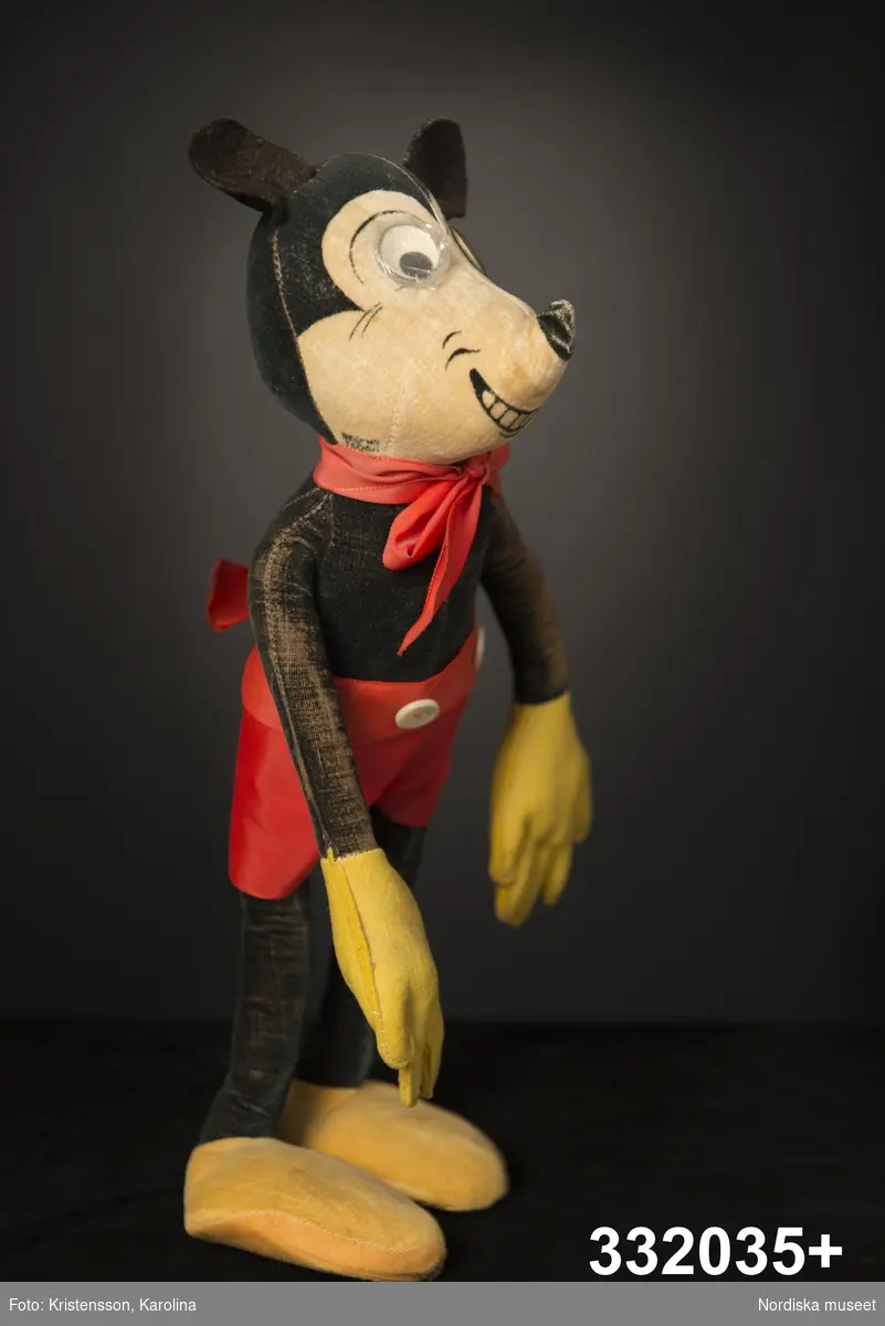 Docka av tyg, föreställande seriefiguren Musse Pigg, en mus klädd i röda kortbyxor och gula handskar och skor.  Lång och smal kropp med böjbara stoppade armar och ben. Stomme av ståltråd, klädd med mjuk stoppning och plysch. Svart kropp och runda musöron av filt, ansikte ljust beige med påklistrade ögon av plast, varav det ena är lagat med tejp och det andra har en spricka. Svart nos, karaktäristiskt leende med synliga tänder målade direkt på tyget. Gula skor och handskar, de senare av filttyg. Tyget är delvis bortnött så att den stålvajer som går genom dockans armar och ben är synlig. Dockan ser ut att vara lagad på några ställen. Det röda bandet med rosett om halsen kan vara ett senare komplement, liksom byxorna med två påsydda vita knappar i linningen.
/Karin Dern 2014-01-27