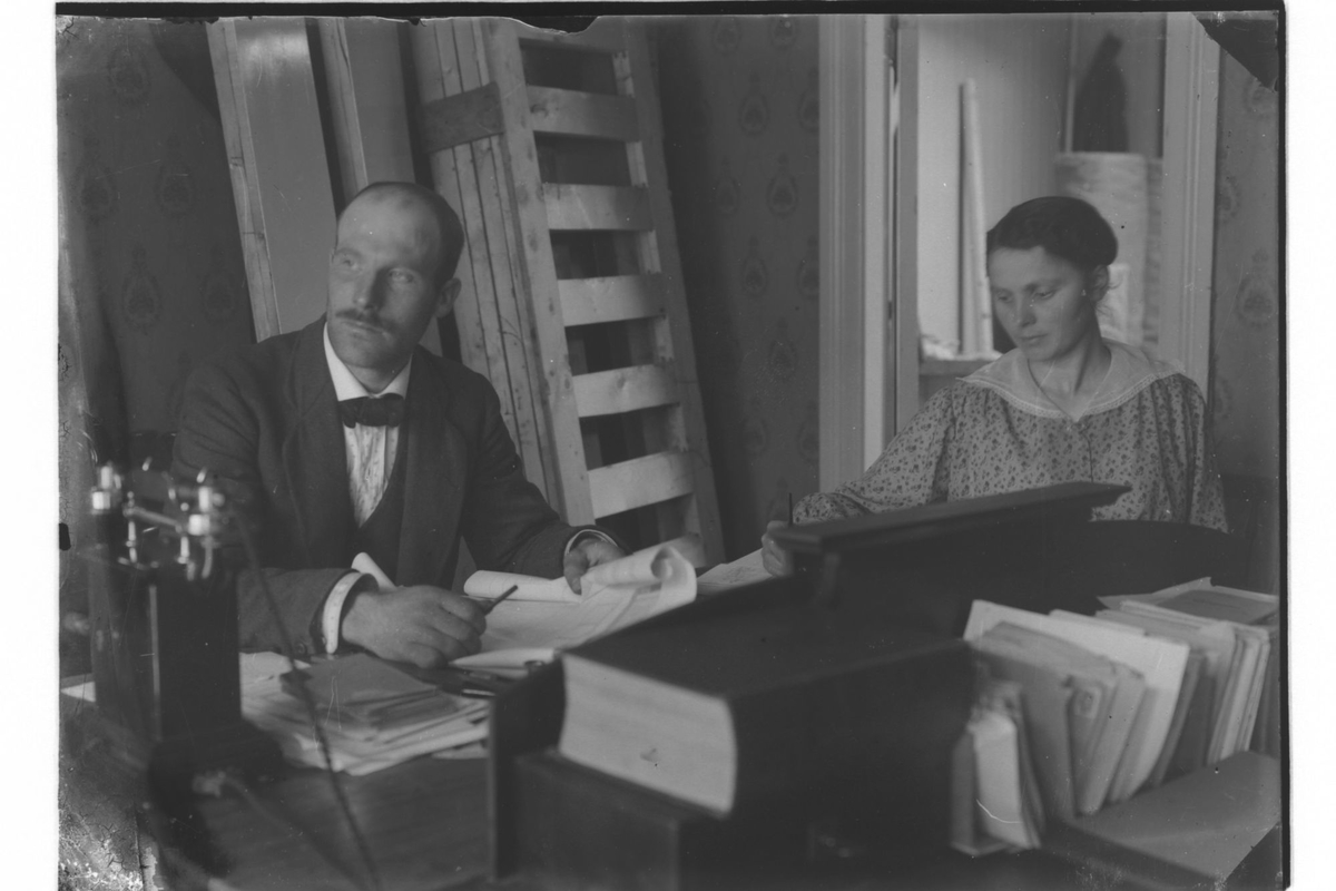 Mann og kvinne ved et skrivebord. Telefonapparat til venstre, og i bakgrunnen står noen sengebunner reist opp mot veggen.