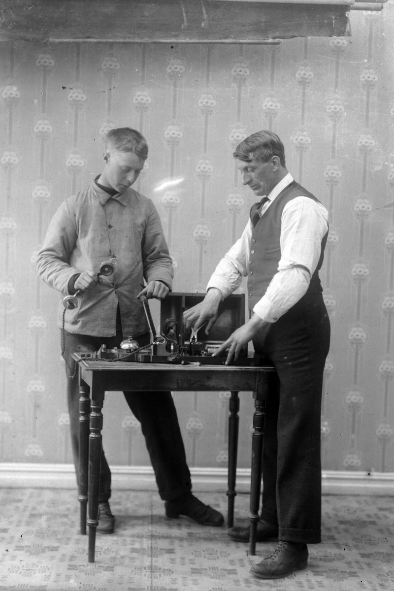 Studioportrett av to menn rundt et bord med telefonapparat på.