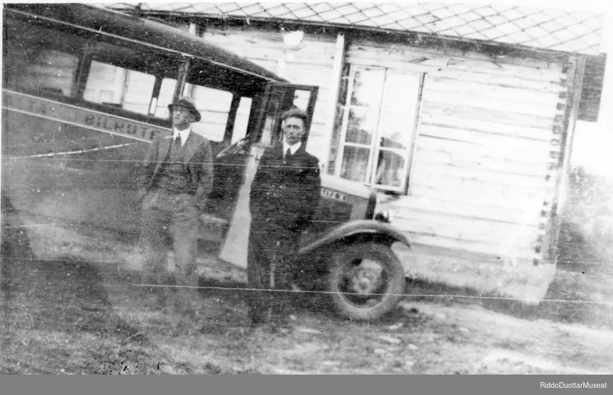 Dievdoolbmot cuožžuba biilla guoras, dimbbarviessu duogabealde.
To menn foran et bil og et tømmer hus i bakgrunnen.