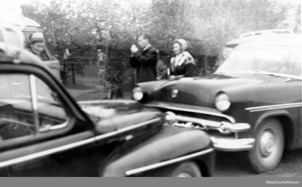 Nissonolbmot ja dievdoolmmoš cužžot guovtti biilla duohken.
Kvinner og en mann står bak to biler.

Volvo PV444, Ford Customline.