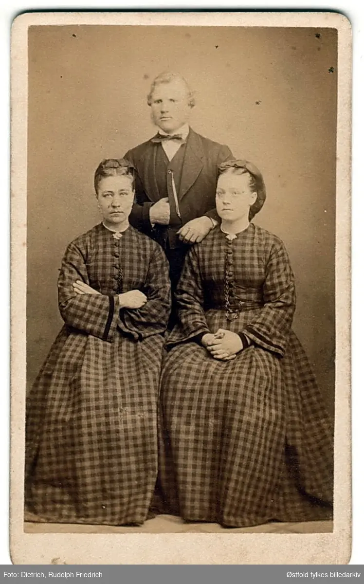To kvinner foran, en mann bak, ukjente, ca. 1865-1881. Firmamerke.
Ukjente personer, ant fra Fredrikstad eller Sarpsborg..