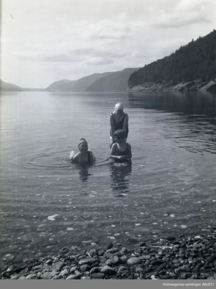 Tre jenter i badetøy på grunna nær land i havet eller en innsjø. I bakgrunnen trær på en åsside. Antatt samme sted og tidspunkt som ST.K.HE 2007-011-0066 til -0071.