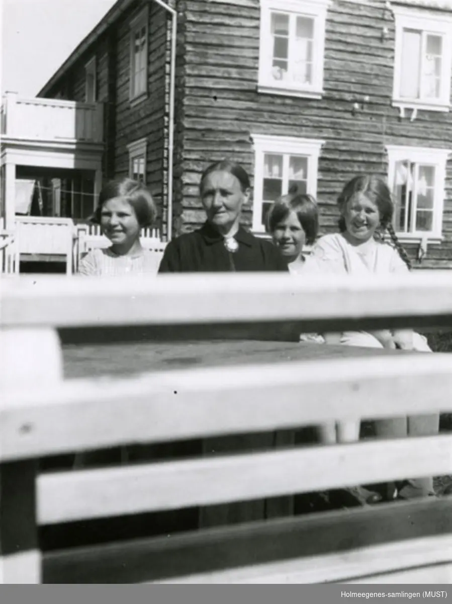En kvinne og tre jenter står foran et tømret hus. Foran dem er et gjerde eller lignende. Se også ST.K.HE 2007-011-0111 til -0126.