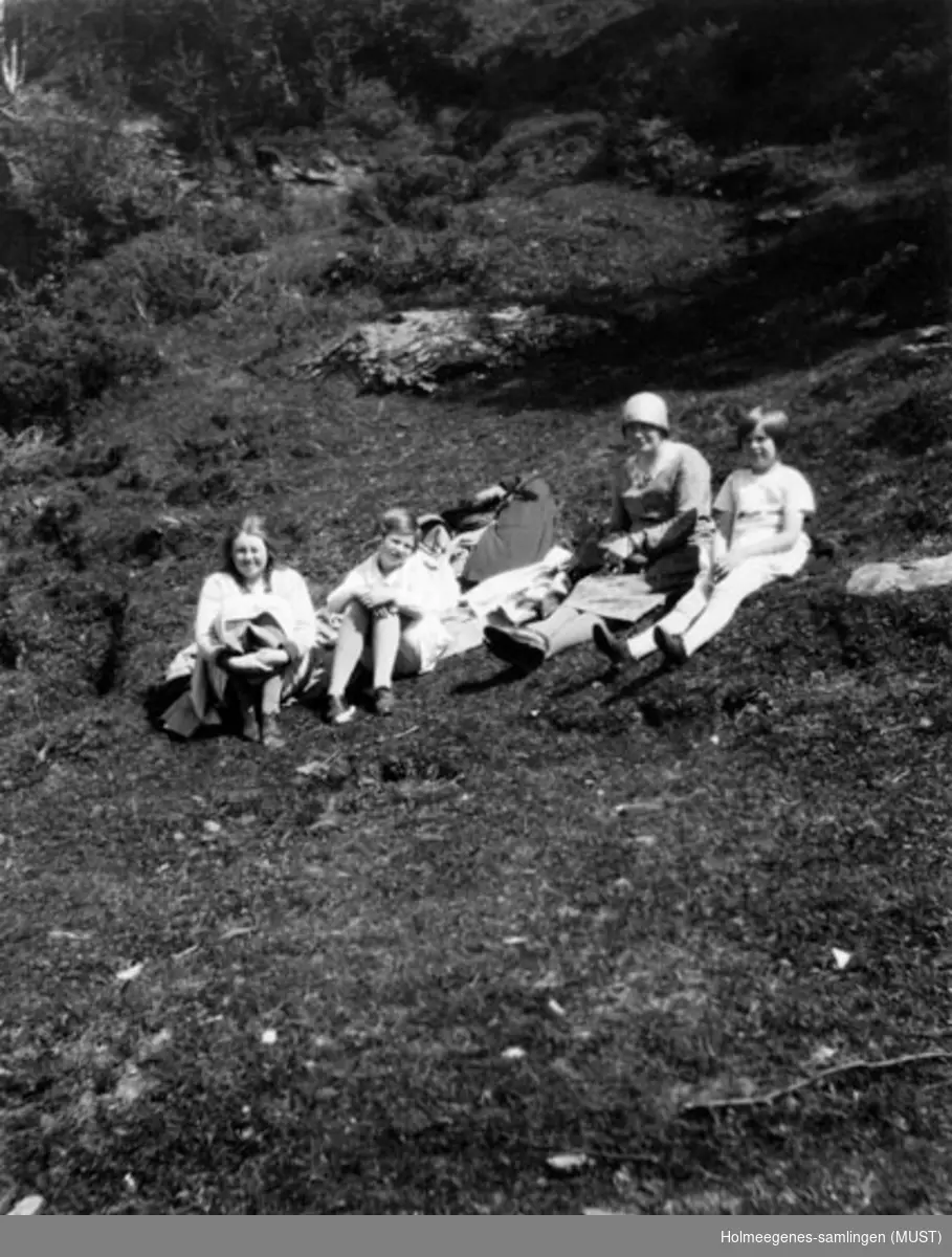 En kvinne og tre jenter i sommerkjoler sitter på pledd el.l. på bakken utendørs. Kvinnen har hatt på seg. Se også ST.K.HE 2007-011-0111 til -0126.