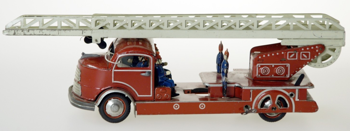 En brandbil, stegbil, av plåt med två brandsoldatfigurer sittande i hytten och fyra på stegflaket. Tillverkad omkring 1950 i U.S.-Zone i Väst-Tyskland.  L. 34 cm.  H. 12 cm.  B 10 cm.