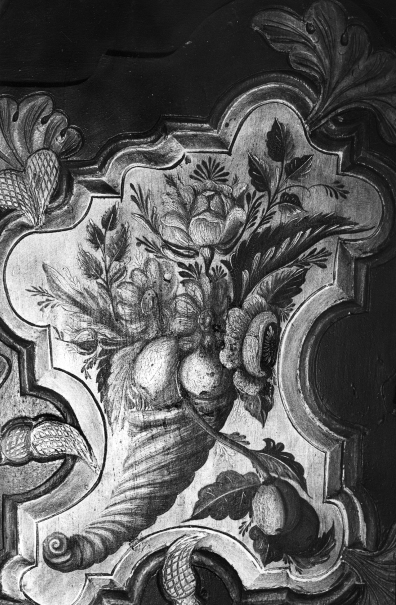 Utskåret med akantusmotiver på framsiden, buet gesims med gjennombrutt akantus og tre oppstikkende palmetter. Skapfeltene malt med blomstermotiv og overflødighetshorns på hvit bunn