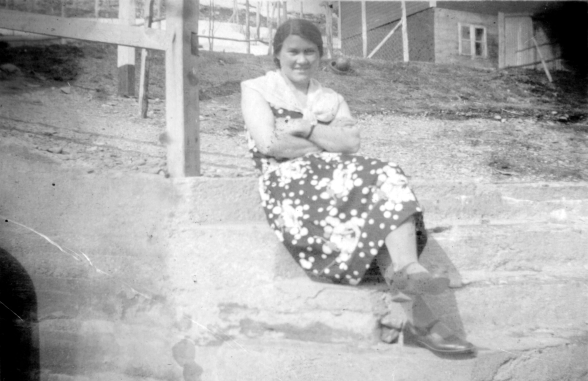 En kvinne sitter på bakken og et hus står i bakgrunnen. Sted og person er ukjent, men bildet kan være tatt i Kvalsund kommune en vårdag, før evakueringa.