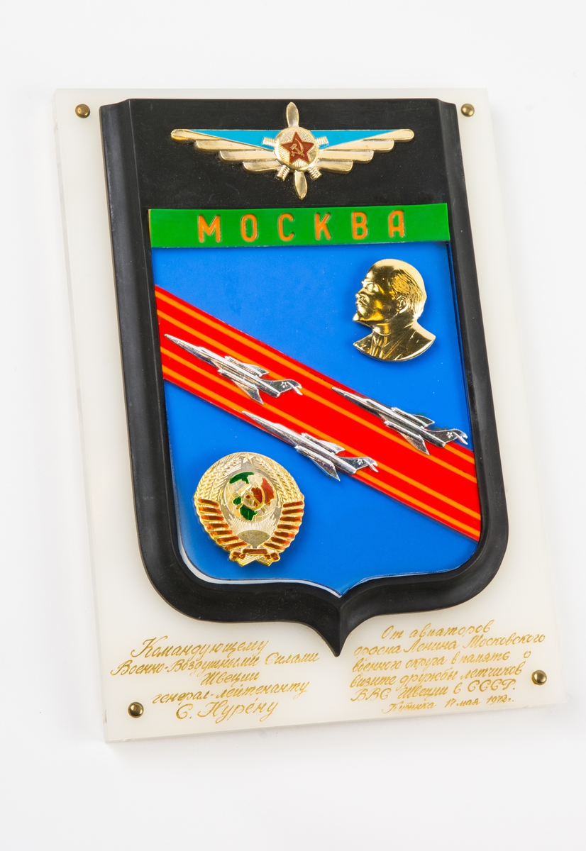 Sköldformad plakett monterad på plastplatta. Plaketten har överst ett flygemblem i guldfärg och blått, i mitten den ryska röda stjärnan med hammaren och skäran. Under detta står MOCKABA med orangefärgade bokstäver på grön botten. Under detta finns på blå botten en siluett i guldfärg föreställande Lenin, snett över plaketten finns ett rött fält med 3 silverfärgade flygplan. Under plaketten en lång text på ryska i guldbokstäver, året är 1972. X = Ett löst runt emblem i guldfärg med motiv av ett rött band, Sädesax, en sol, en jordglob och hammaren och skäran.