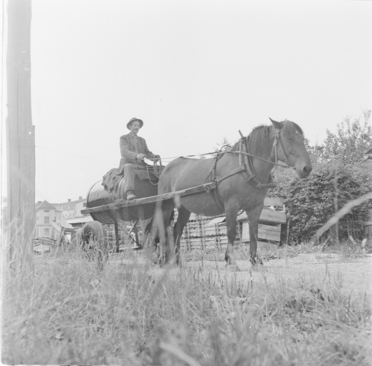 Man, möjligen Axel Andersson, Kvek, med häst och vagn, gamla brännvinsbränneriet i bakgrunden, Örsundsbro, Gryta socken, Uppland 1955