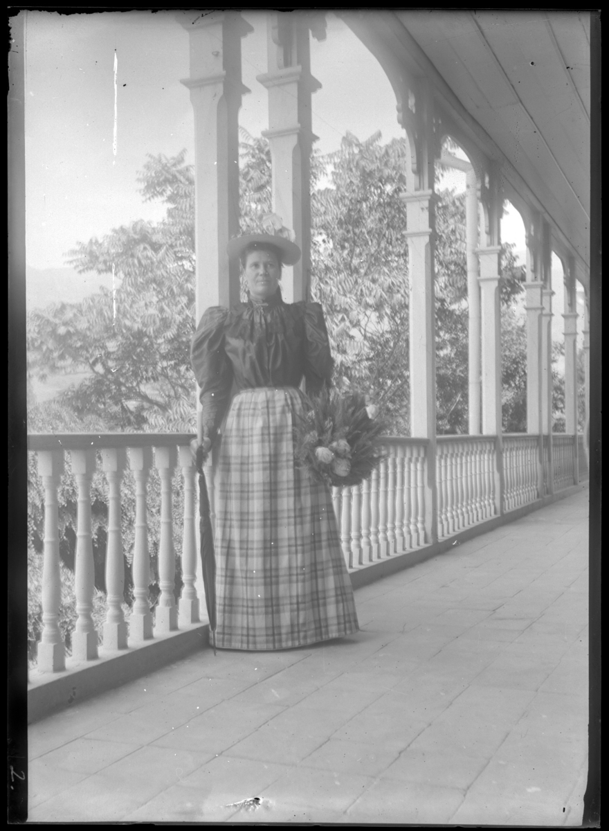 '1 kvinna stående på balkong med trägolv och träräcke. Kläder. Klädsel. Hatt. Blombukett i handen. ::  :: Ingår i serie med fotonr. 5280:1-17. Se även fotonr. 5269-5292 med bilder från Stuxbergs resa till Kaukasien.'