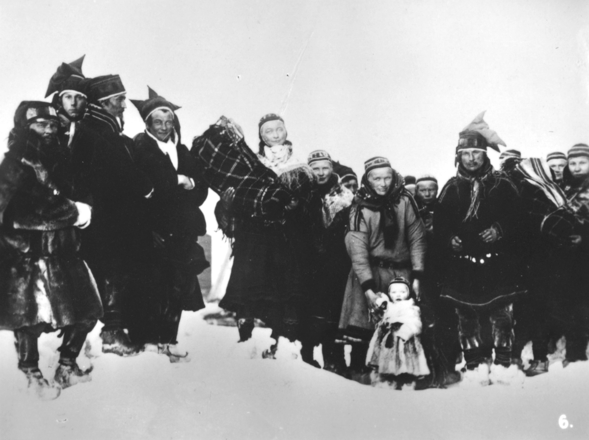 Bilde nr. 6 i serien '10 amatørbilleder fra lappernes hjem og liv i Finnmark', se FB 93164-001. 'Lappebarna bringes til kirke for at døpes.'  En gruppe samer kledt i pesker og kofter. Menn med stjerneluer, kvinner i luer og sjal. Skaller på beina. To barn ligger i komse, et barn står foran, kledt i pesk.